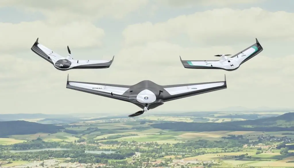 três drones de asa fixa, conhecidos como VANTs (Veículos Aéreos Não Tripulados), em pleno voo sobre uma vasta