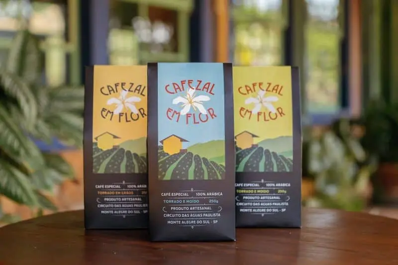 três embalagens de café da marca ‘Cafezal em Flor’, posicionadas verticalmente sobre uma superfície de madeira