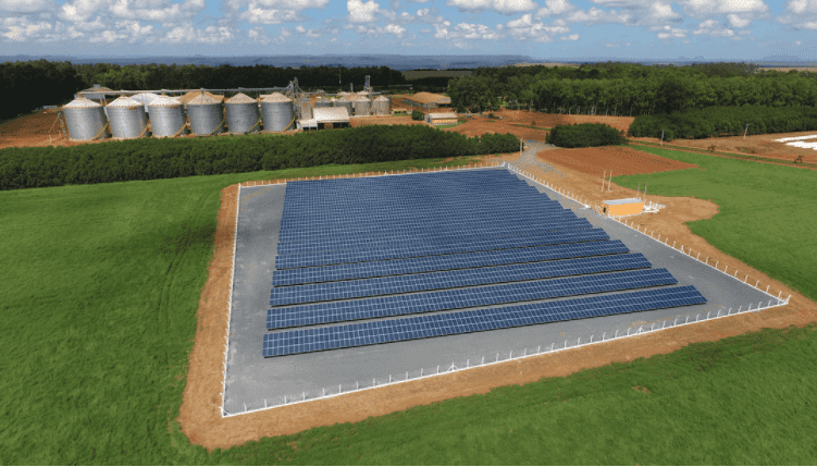 uma vista aérea de uma grande instalação de painéis solares em uma propriedade rural. Em primeiro plano, cente