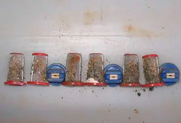 uma fileira de filtros de bico de um pulverizador agrícola, visivelmente sujos e obstruídos. São sete filtros
