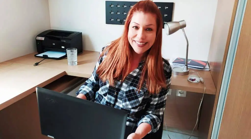 uma mulher sorridente, com cabelos ruivos e vestindo uma camisa xadrez, sentada em seu escritório. Ela segura