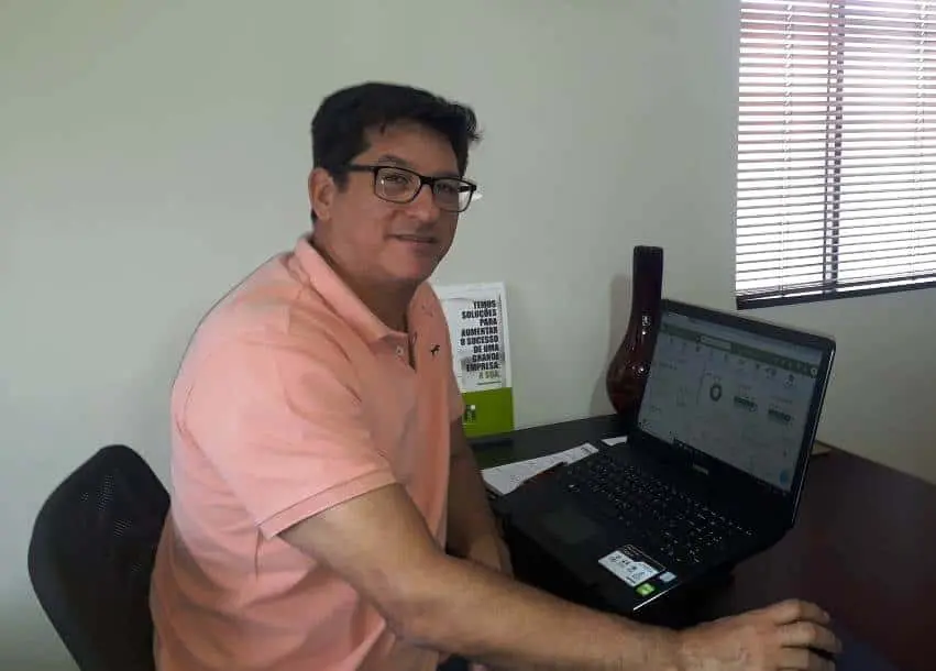 homem de meia-idade, com óculos e camisa polo de cor salmão, sentado em sua mesa de trabalho em um ambien