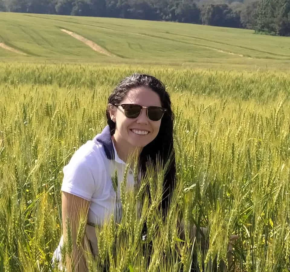 mulher sorridente, com cabelos escuros e óculos de sol, em meio a uma vasta plantação de trigo. Ela vest