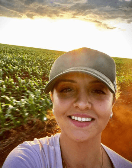 selfie tirada por uma mulher jovem e sorridente, que usa um boné. Ela está em uma grande lavoura, possivelment