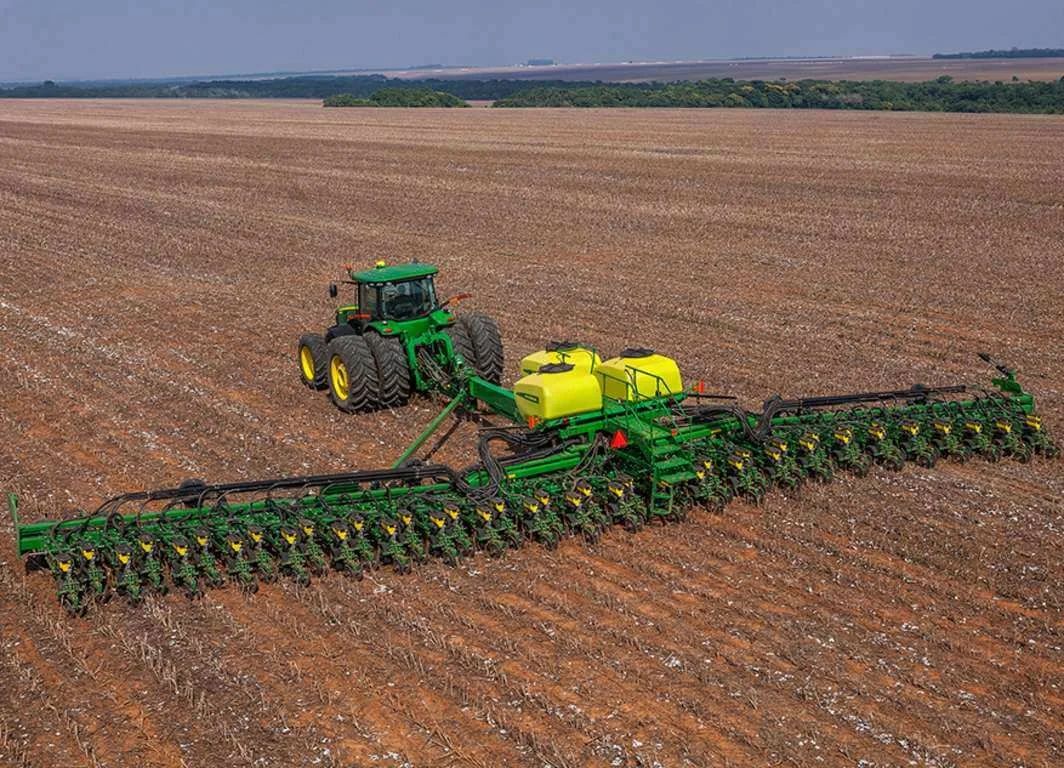 uma operação agrícola de grande escala, capturada de um ângulo elevado. No centro, um trator verde da marca Jo