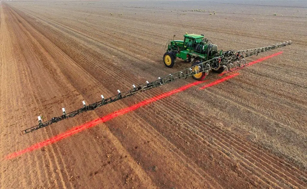 uma visão aérea de um pulverizador agrícola de grande porte, acoplado a um trator verde, operando em uma vasta