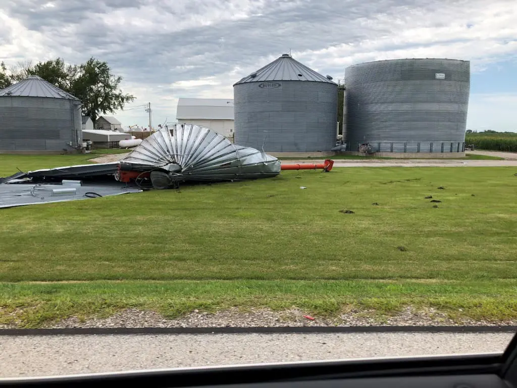 o cenário de uma propriedade rural após um evento climático severo. Em primeiro plano, um grande silo de grã