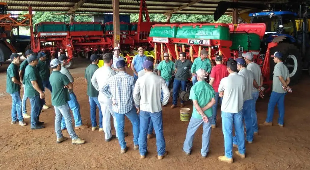 grupo de agricultores e operadores de máquinas reunidos em um grande galpão de fazenda, com piso de terra